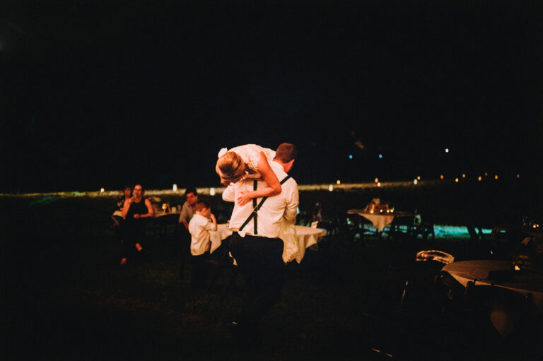 A groom carries his bride over his shoulder late at night during an outdoor wedding reception. Guests sit at candlelit tables in the background while laughter and motion fill the frame. Captured by San Antonio documentary wedding photographer Christian Margain.